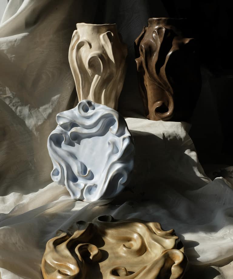 Four sculpted ceramic vessels in white and brown tones, featuring organic flowing forms, arranged on draped fabric.