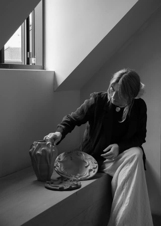 Fanny Schultz seated on a window bench, reaching for the Floral Vase beside two glazed platters