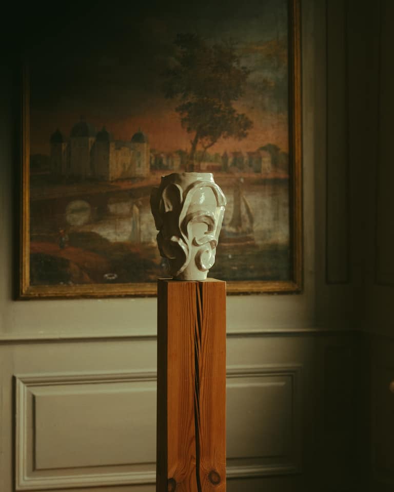A white sculpted vase sits on a wooden pedestal in front of an oil painting depicting a landscape with a castle.